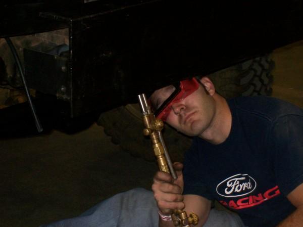 Man in Ford Racing shirt and red safety glasses holding a welding torch under a black vehicle frame.