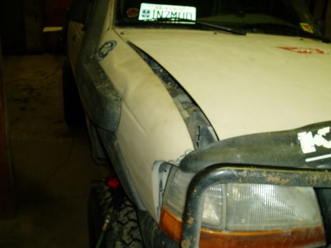 2000 Ford E350 - A close-up, low-angle shot of the front passenger side of a white vehicle, possibly a van or SUV, showing damage to the fender and hood. A…