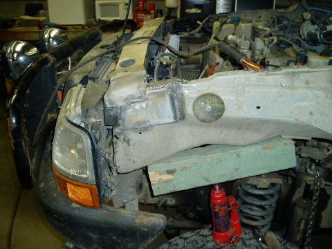 2000 Ford E350 - A close-up view of the front passenger side of a damaged vehicle, showing a broken headlight, bent metal bodywork, and engine components. A…
