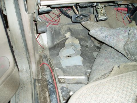 2000 Ford E350 - The interior floor of a vehicle shows exposed wiring and a damaged metal floor panel near the pedals. A gray fabric mat is partially coveri…