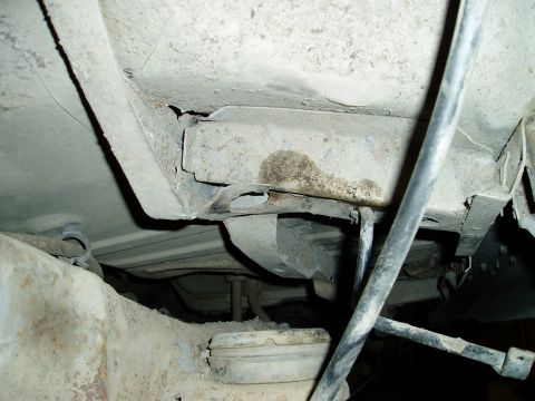 2000 Ford E350 - The underside of a vehicle shows rusted metal framing and suspension components, with a dark cable running diagonally across the frame.