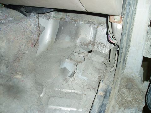 2000 Ford E350 - The interior floor of a vehicle shows significant wear and tear, with exposed metal, torn carpeting, and visible wiring. The area appears t…