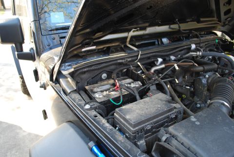 2008 Jeep Wrangler - The open hood of a black Jeep Wrangler reveals the engine bay, focusing on the battery and fuse box. The vehicle is parked outdoors on a pa…
