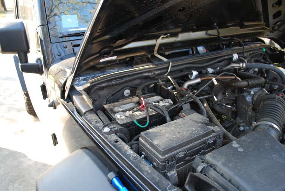 Black Jeep Wrangler engine bay with battery and fuse box visible under the open hood.