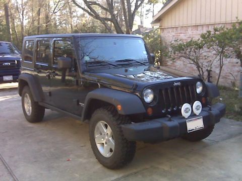 2008 Jeep Wrangler - A black four-door Jeep Wrangler is parked on a concrete driveway in front of a brick house. The Jeep has aftermarket fog lights mounted on…
