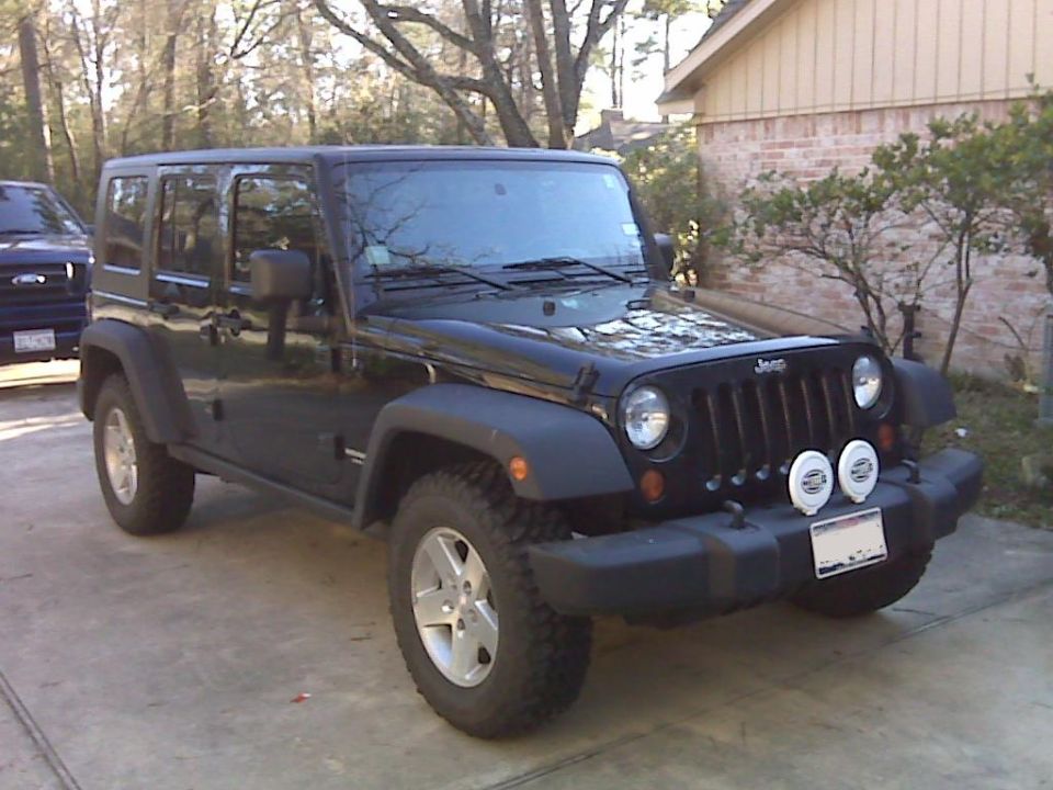 Black four-door Jeep Wrangler with aftermarket fog lights parked on a driveway.