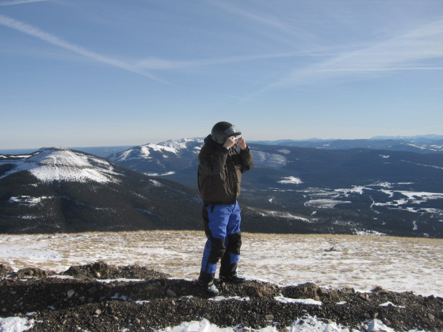 Person in helmet and winter gear on snowy hilltop overlooking mountains.