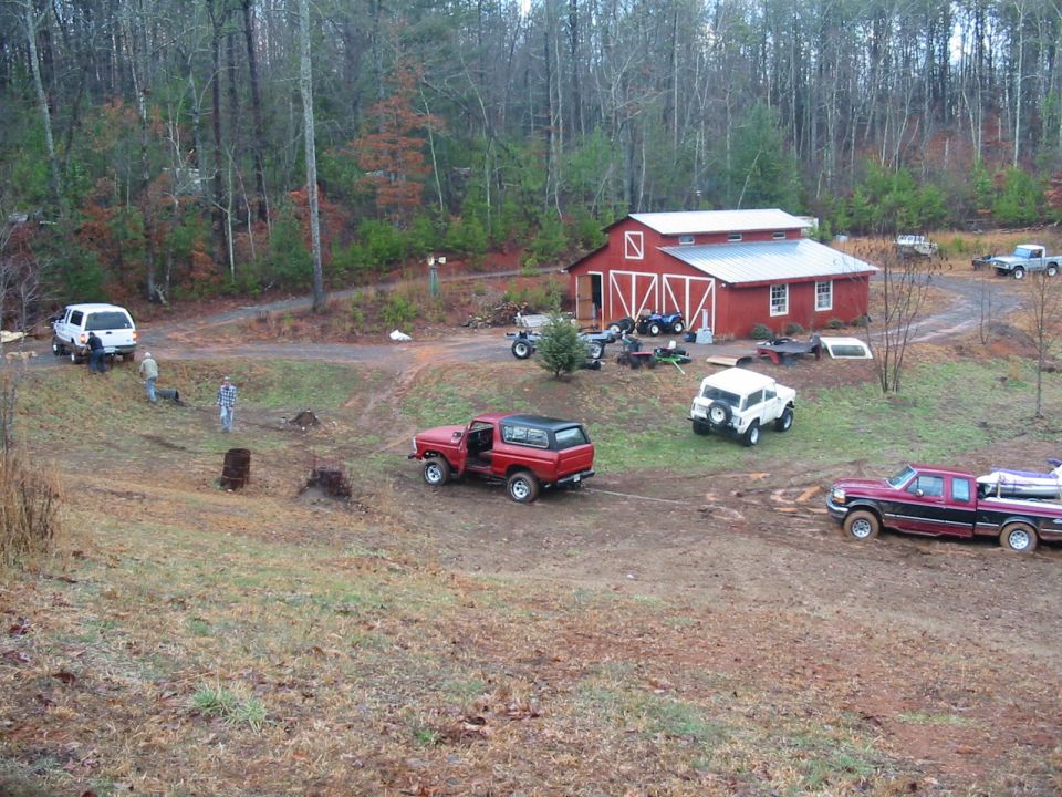 Red Ford Bronco with door open in muddy field near red barn, with other trucks and ATVs nearby