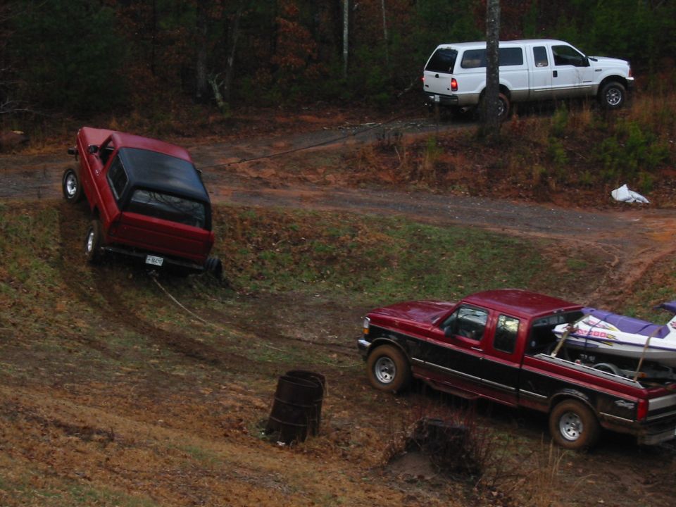 Red Bronco stuck on muddy hillside being tow-strapped out by red F-150 and white Ford pickup.