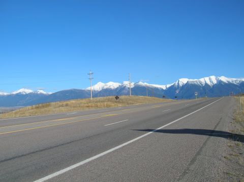 1996 Ford Bronco - A paved highway curves towards snow-capped mountains under a clear blue sky. Telephone poles line the side of the road, and dry grass cover…