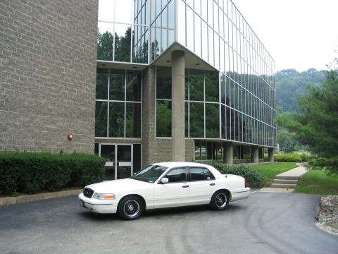 1998 Ford Crown Victoria - A white 1998 Ford Crown Victoria is parked on an asphalt driveway in front of a building with reflective glass windows.