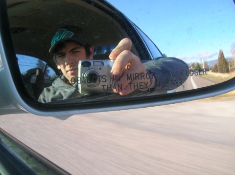 1996 Ford Bronco - A young man wearing a baseball cap takes a selfie in a car's side mirror, holding a digital camera. The mirror reflects the driver and the…