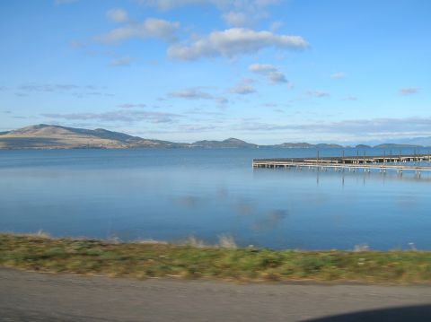 1996 Ford Bronco - A wide shot of a calm lake with mountains in the distance and a wooden pier extending into the water. The foreground shows a grassy embankm…