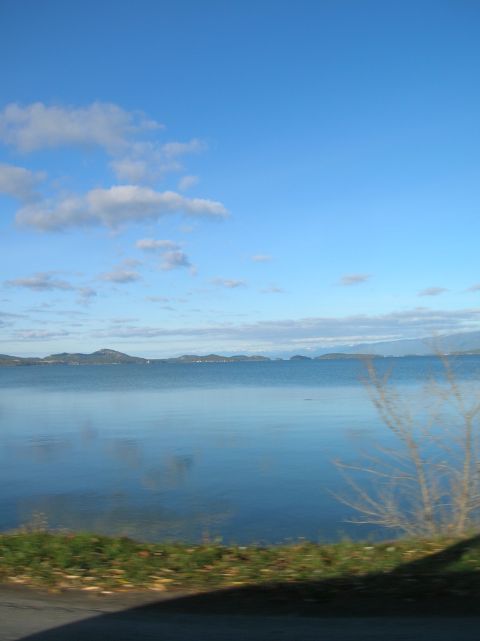 1996 Ford Bronco - A wide shot of a calm lake with distant islands under a bright blue sky with scattered clouds. The foreground shows a grassy embankment wit…