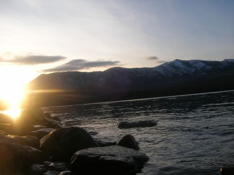 1996 Ford Bronco - The sun sets behind snow-capped mountains, casting a warm glow over a rocky shoreline and calm lake.