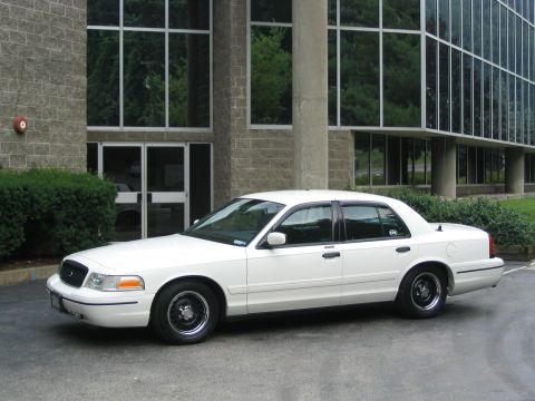 1998 Ford Crown Victoria - A white 1998 Ford Crown Victoria sedan is seen from the side, parked in front of a building with large windows.