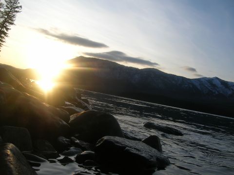 1996 Ford Bronco - The sun sets behind a mountain range, casting a warm glow over a rocky shoreline and a body of water.