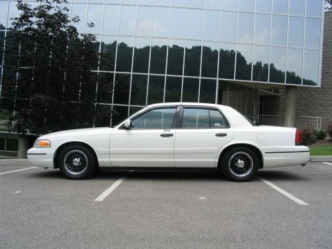 1998 Ford Crown Victoria - A white 1998 Ford Crown Victoria sedan is parked in a lot in front of a modern building.