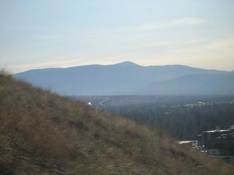 1996 Ford Bronco - A wide shot of a hazy mountain range in the background, with a town nestled in the valley below. In the foreground, dry grass covers a stee…