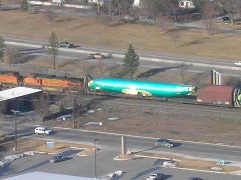1996 Ford Bronco - An aerial view shows a train carrying a large, green, cylindrical fuselage, likely for an aircraft, on a flatbed car. The train is moving a…