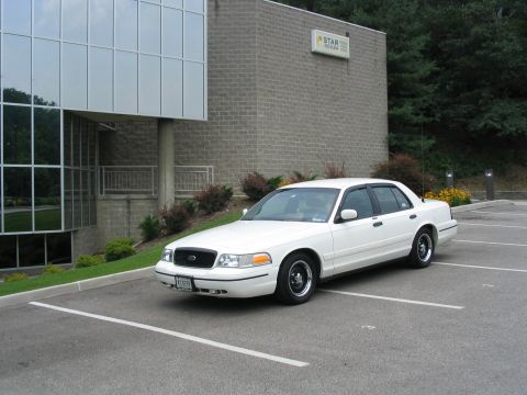 1998 Ford Crown Victoria - A white 1998 Ford Crown Victoria is parked in a lot next to a modern building.