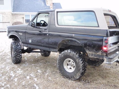 1996 Ford Bronco - A lifted black 1996 Ford Bronco with large off-road tires is parked on a grassy area with patches of snow. The vehicle is viewed from the s…