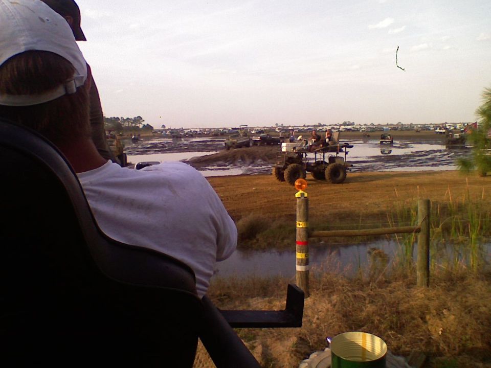 Custom off-road vehicle with large tires driving through mud and water with spectators in the background.