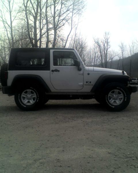 2005 Jeep Wrangler - Side profile of a white 2-door Jeep Wrangler with a black hardtop parked on a dirt road. The vehicle has black fender flares and BFGoodrich…