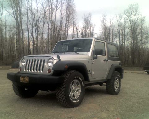 2005 Jeep Wrangler - A silver 2-door Jeep Wrangler is parked on a gravel lot with a bare-branched forest in the background. The Jeep has a black hardtop and aft…