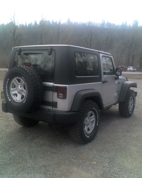 2005 Jeep Wrangler - The rear passenger side of a silver 2-door Jeep Wrangler is shown parked on a gravel surface with a wooded hillside in the background. The…