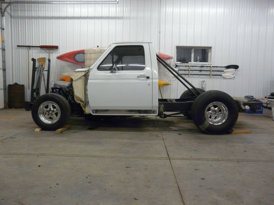 Side profile of a white Ford pickup cab on a bare frame with tube brace inside a shop