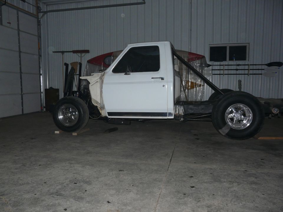 White Ford pickup cab on bare frame with tube bracing and polished wheels inside a shop