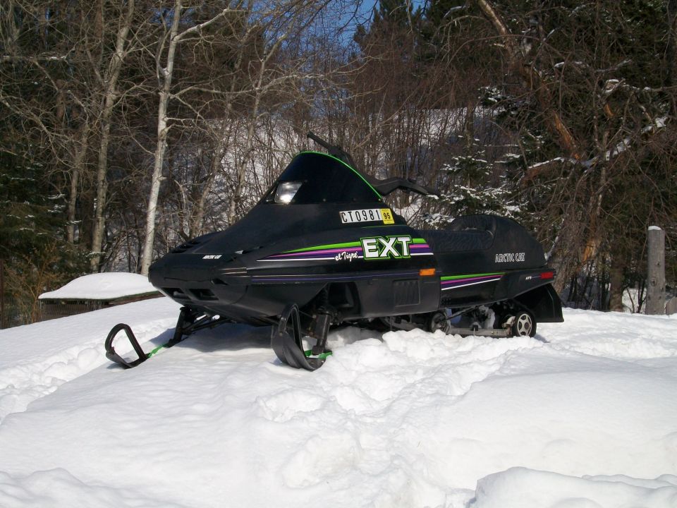 Black Arctic Cat EXT snowmobile parked on snow among bare trees.