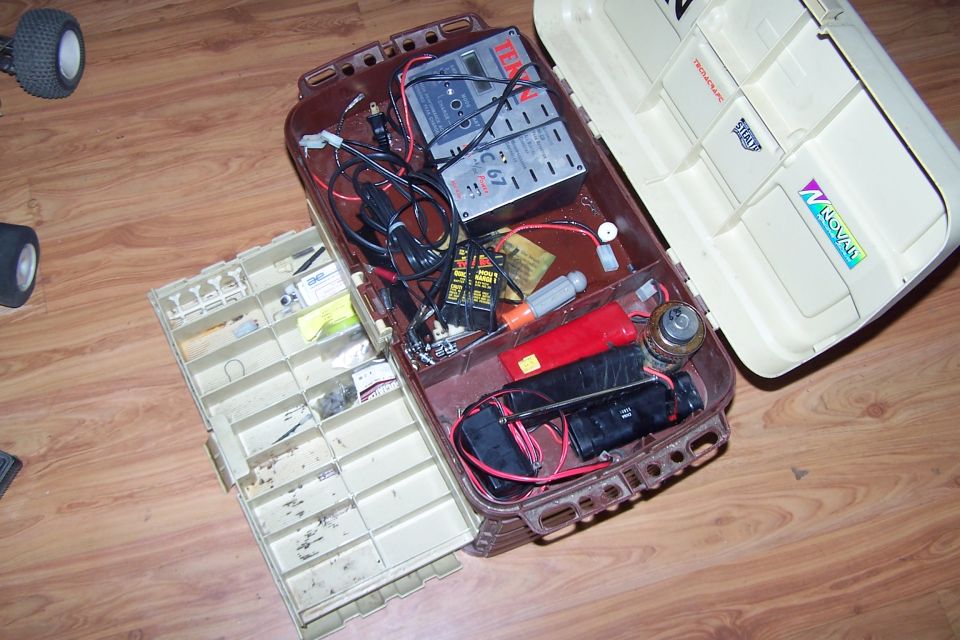 Open tool box with RC charger, wires, batteries and parts tray on wood floor