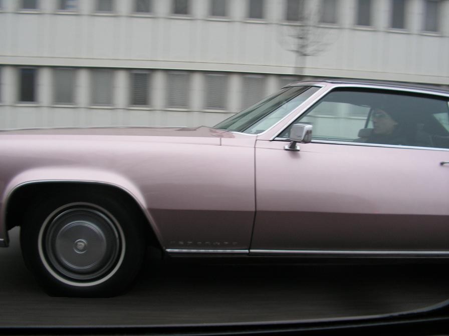 Side view of a pink Cadillac Eldorado driving past a building