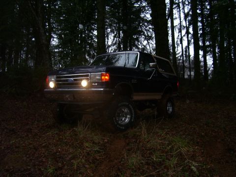 1991 Ford Bronco - A dark Ford Bronco navigates a wooded area with its headlights on.