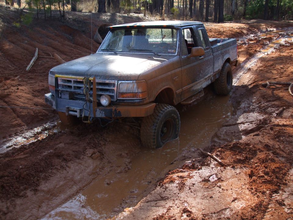 Muddy lifted Ford F-150 extended cab pickup in deep water-filled ruts on a red clay trail