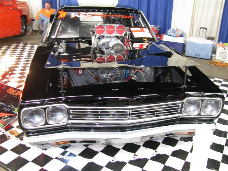 Black custom car with a large supercharger and hood open, on a checkered floor at a show