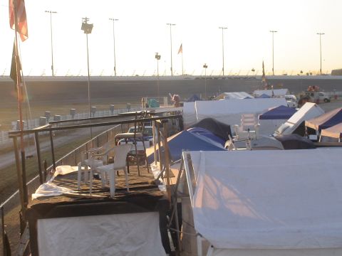 1993 Ford C-350 (Centurion Conversions) - A campsite is set up adjacent to a race track, with tents and some vehicles under a bright sky.