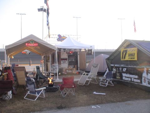 1993 Ford C-350 (Centurion Conversions) - Campsites are set up at a race track, tents, chairs, a fire pit, and various sponsor banners.