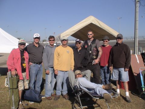 1993 Ford C-350 (Centurion Conversions) - A group of men stand and recline outdoors, with tents and a race track visible in the background under a clear blue sky.