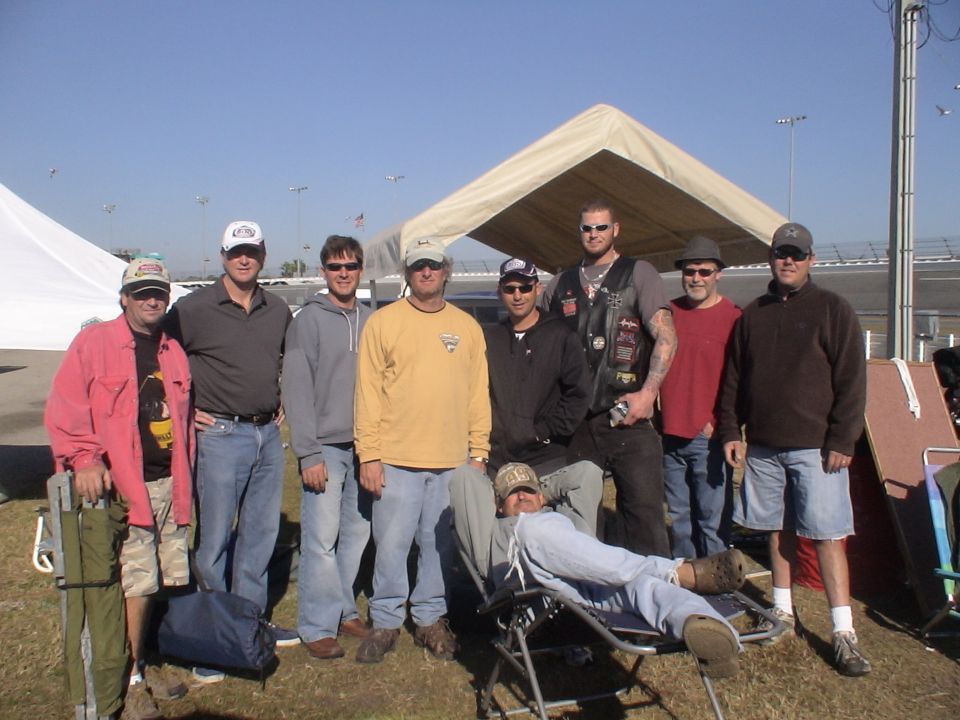 Group of men posing outdoors with tents and a race track in the background