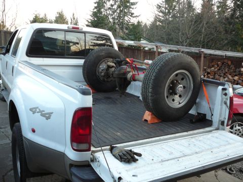 1991 Ford Bronco - A white Ford F-Series truck carries a rear axle and tires in its bed, with a red car visible to the right.