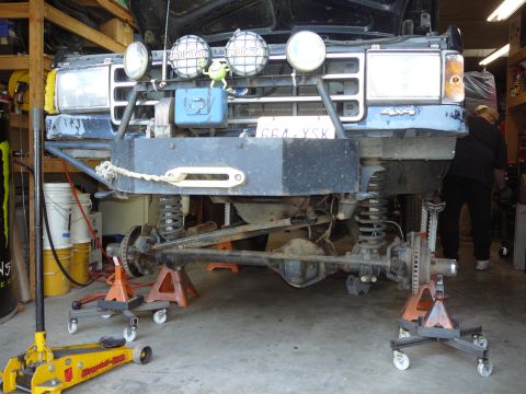 1991 Ford Bronco - The front axle and suspension of a Ford Bronco are visible as the vehicle is raised on jack stands in a garage.