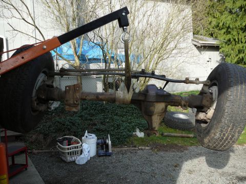 1991 Ford Bronco - A Ford Bronco front axle assembly is being lifted by an engine hoist, showing its heavy-duty components and large tires.