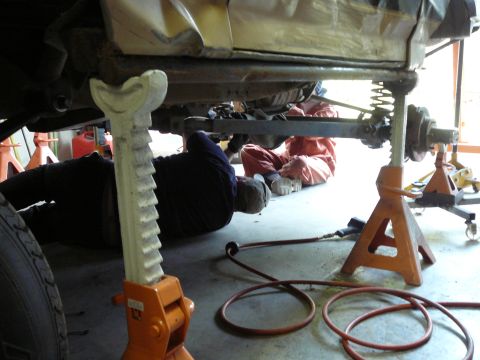 1991 Ford Bronco - Two mechanics are seen working on the underside of a vehicle that is supported by jack stands in a garage.