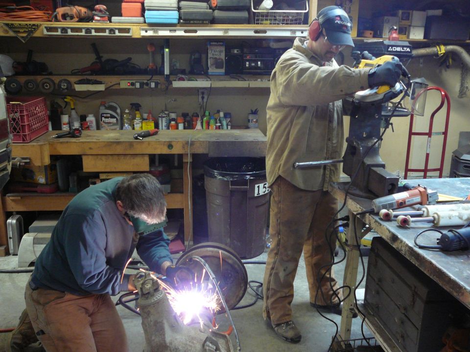 Two men welding and grinding in a cluttered garage workshop with tools and shelves