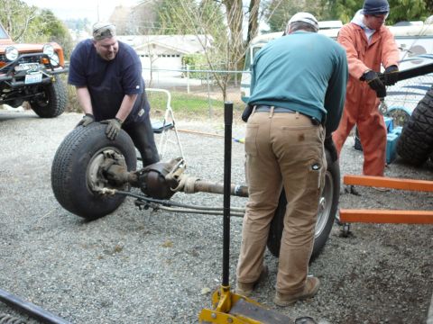 1991 Ford Bronco - Men are handling a vehicle axle and wheel assembly, with another vehicle in the background.
