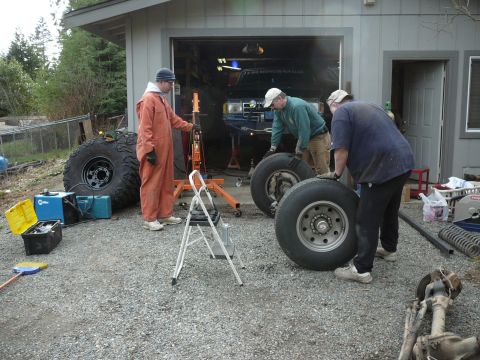 1991 Ford Bronco - A group of people are gathered around a vehicle, preparing to change its large tires in front of a garage.