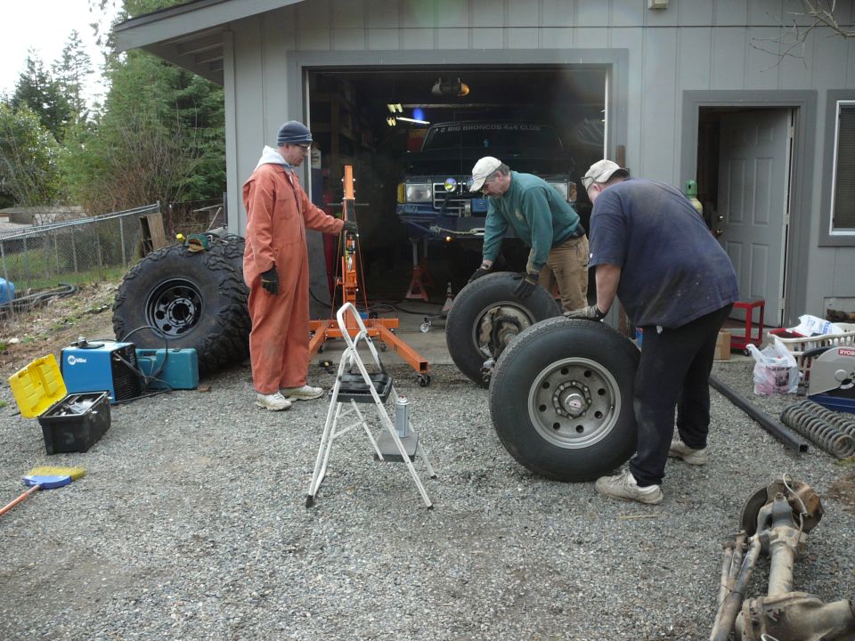 Three guys are working on a vehicle in front of a garage. Two are moving large tires, one is standing nearby. A blue truck is visible inside the garage